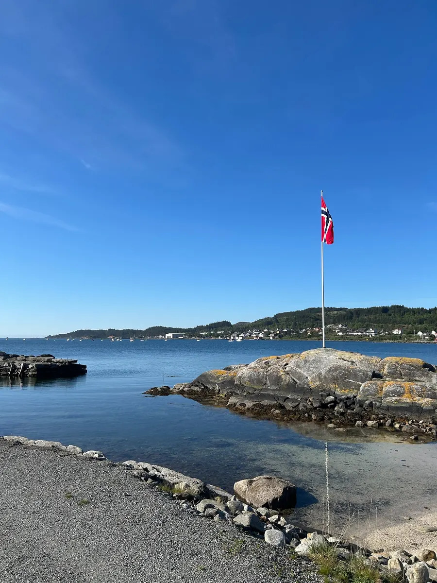 Svaberg og klart vann ved campingplassen, med flaggstang mot blå himmel